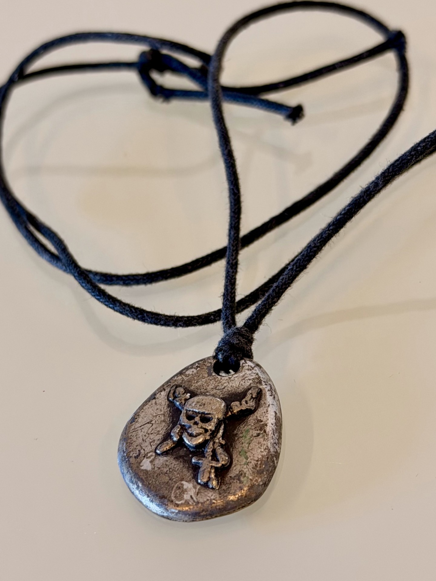 Un teschio da portare al collo: ciondolo ovale con teschio e ossa in rilievo, finitura color argento invecchiato e cordino scuro, dalla raccolta di Alfnet.